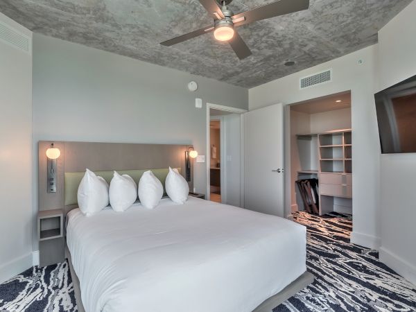 Modern bedroom with a large bed, five pillows, nightstands, ceiling fan, wall-mounted TV, closet area, and patterned carpet.