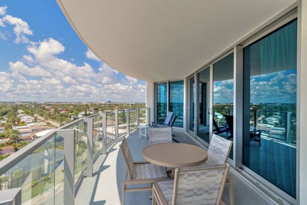 A balcony with glass railing features outdoor seating, a table, and a scenic view of a city skyline and blue sky with clouds.