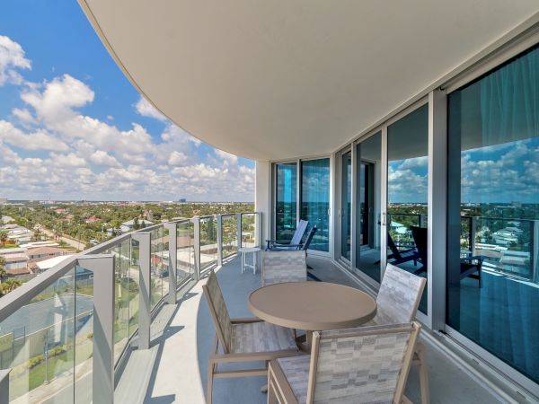 A balcony with glass railing features outdoor seating, a table, and a scenic view of a city skyline and blue sky with clouds.