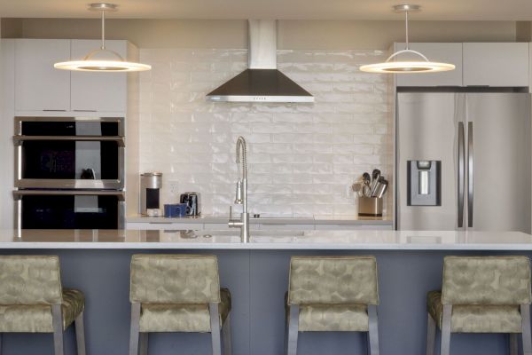 A modern kitchen featuring a central island with four chairs, stainless steel appliances, a double oven, hanging lights, and a sleek backsplash.