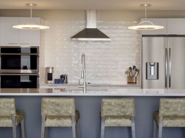 A modern kitchen featuring a central island with four chairs, stainless steel appliances, a double oven, hanging lights, and a sleek backsplash.