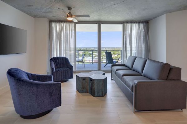 A modern living room with a grey couch, two blue chairs, a hexagonal coffee table, and a wall-mounted TV. It has a large window with a view.