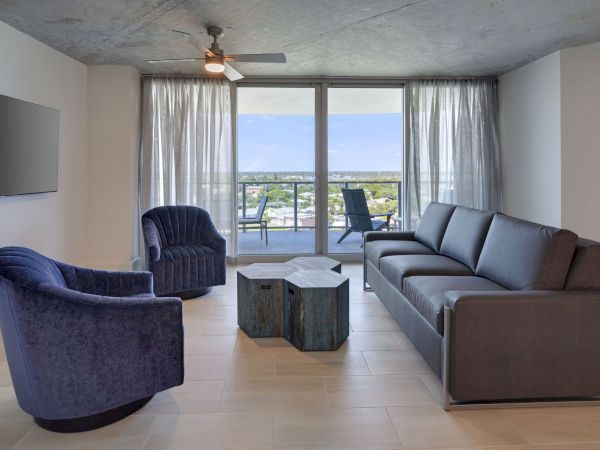 A modern living room with a grey couch, two blue chairs, a hexagonal coffee table, and a wall-mounted TV. It has a large window with a view.