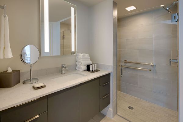 A modern bathroom with a wide countertop, mirror, magnifying mirror, soap dish, and towels. A shower space with a glass partition is visible.