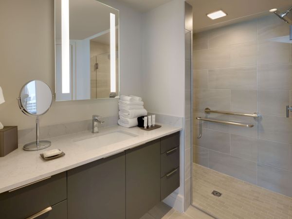 A modern bathroom with a wide countertop, mirror, magnifying mirror, soap dish, and towels. A shower space with a glass partition is visible.
