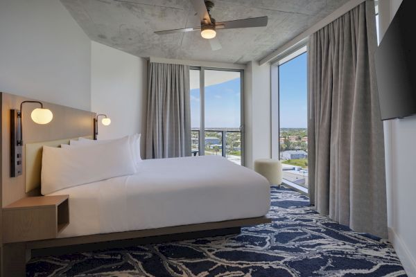 The image shows a modern hotel room with a large bed, night lamps, a ceiling fan, a large window with a city view, and a patterned blue carpet.