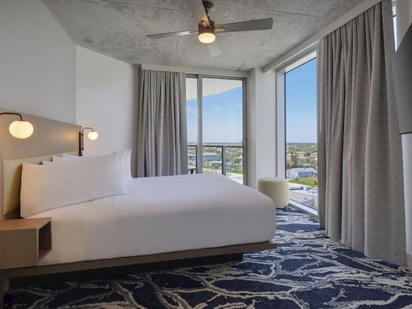 The image shows a modern hotel room with a large bed, night lamps, a ceiling fan, a large window with a city view, and a patterned blue carpet.