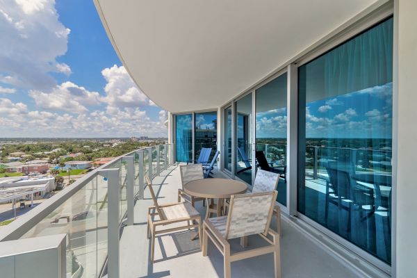 This image shows a modern balcony with glass railings and outdoor furniture, overlooking a cityscape under a blue sky with scattered clouds.