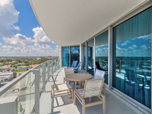 This image shows a modern balcony with glass railings and outdoor furniture, overlooking a cityscape under a blue sky with scattered clouds.