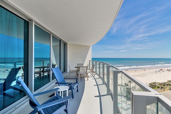 A modern balcony with blue chairs and a table overlooks a serene beach and the ocean under a clear blue sky, perfect for relaxation.