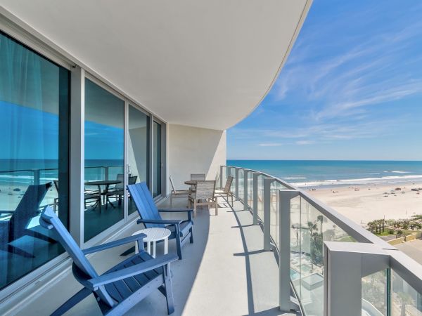A modern balcony with blue chairs and a table overlooks a serene beach and the ocean under a clear blue sky, perfect for relaxation.