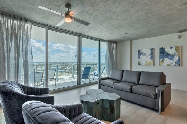 A modern living room with large windows, two chairs, a couch, coffee table, ceiling fan, and balcony with outdoor furniture, offering an ocean view.