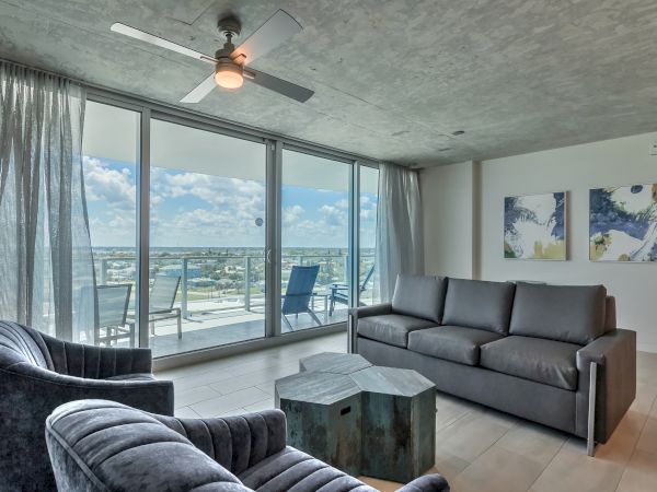 A modern living room with large windows, two chairs, a couch, coffee table, ceiling fan, and balcony with outdoor furniture, offering an ocean view.