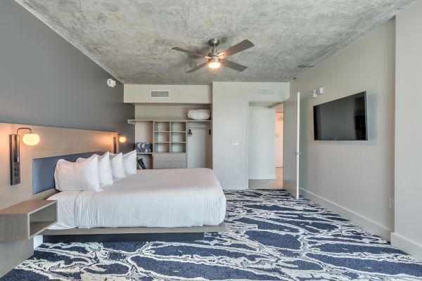 A modern bedroom with a double bed, multiple pillows, wall-mounted TV, ceiling fan, open closet, and patterned blue carpet.