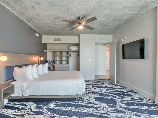 A modern bedroom with a double bed, multiple pillows, wall-mounted TV, ceiling fan, open closet, and patterned blue carpet.