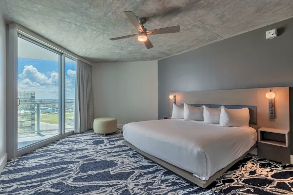 This image shows a modern bedroom with a large bed, patterned carpet, ceiling fan, and floor-to-ceiling window offering a balcony view.