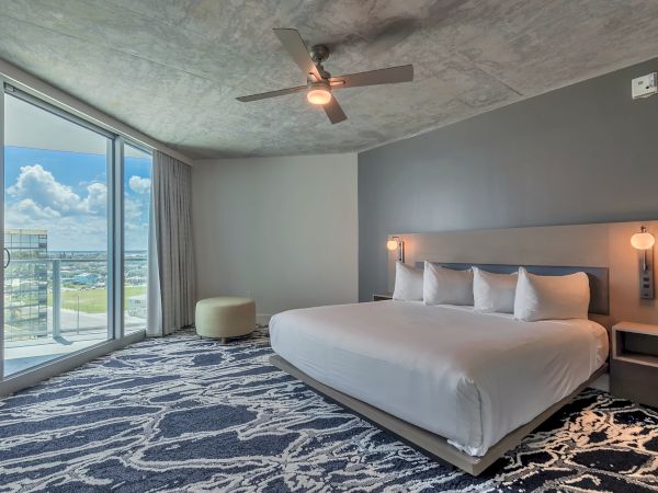 This image shows a modern bedroom with a large bed, patterned carpet, ceiling fan, and floor-to-ceiling window offering a balcony view.