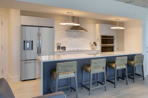 Modern kitchen with stainless steel appliances, including a refrigerator and oven, a white island countertop, and four green upholstered bar stools.