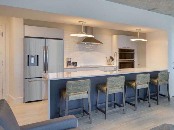 Modern kitchen with stainless steel appliances, including a refrigerator and oven, a white island countertop, and four green upholstered bar stools.