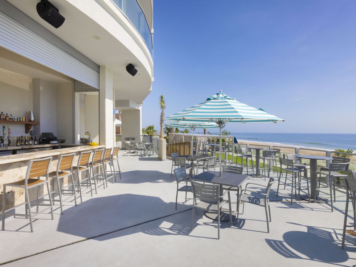 An outdoor beachfront bar with seating, tables, and a striped umbrella overlooking the ocean under a clear blue sky.