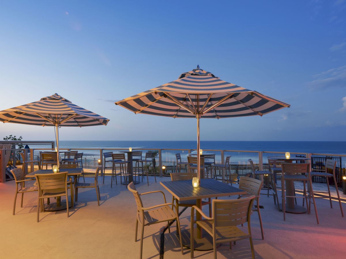 An outdoor cafe with wooden tables, chairs, and striped umbrellas by the sea during sunset, offering a serene view of the ocean and used candles.