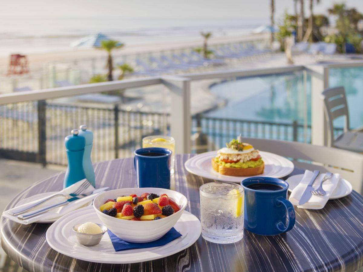 A breakfast table with a bowl of fruit, coffee, water, and a dish topped with various foods, set beside a pool with an ocean view.