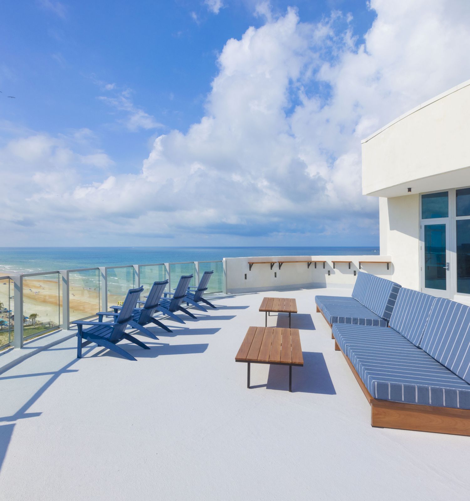 A rooftop terrace with ocean view, blue chairs, a striped cushioned bench, and tables, bordered by glass panels and a modern building.