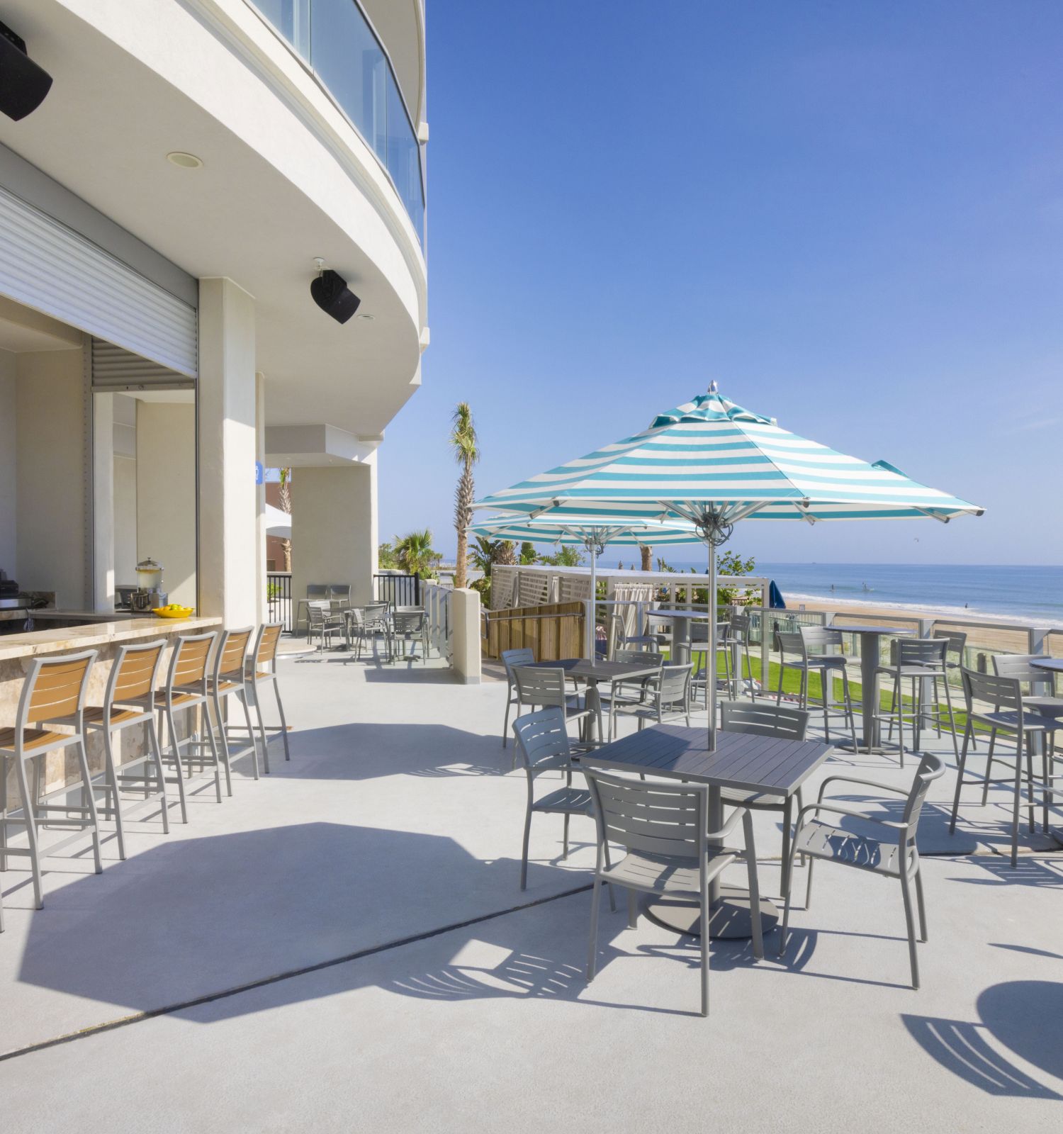 An outdoor beachside bar with stools and tables, shaded by umbrellas, overlooking the ocean on a sunny day, creating a relaxing atmosphere.