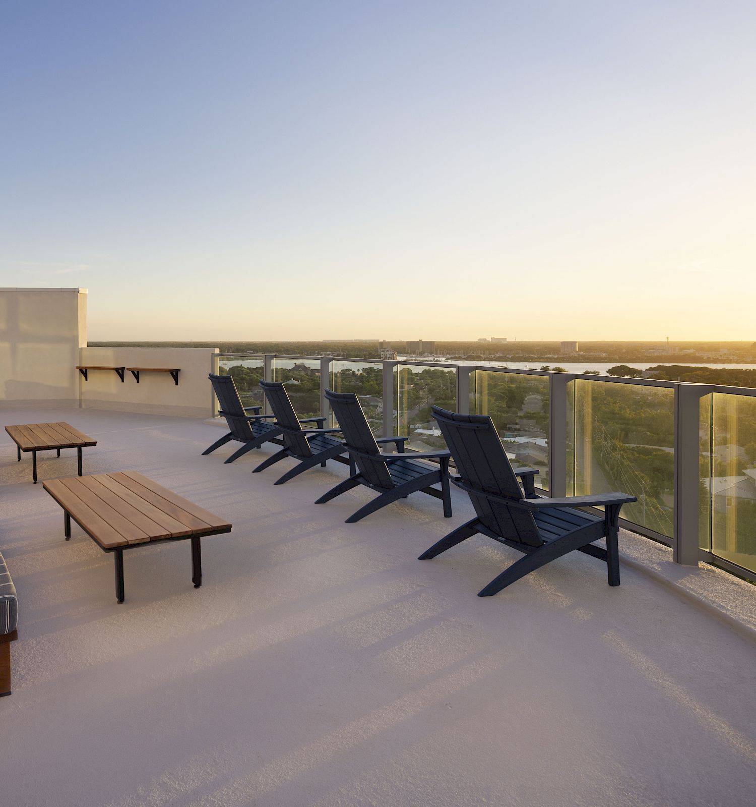 The image shows a rooftop terrace with lounge chairs and tables, a glass safety railing, and a view of the horizon at sunset.