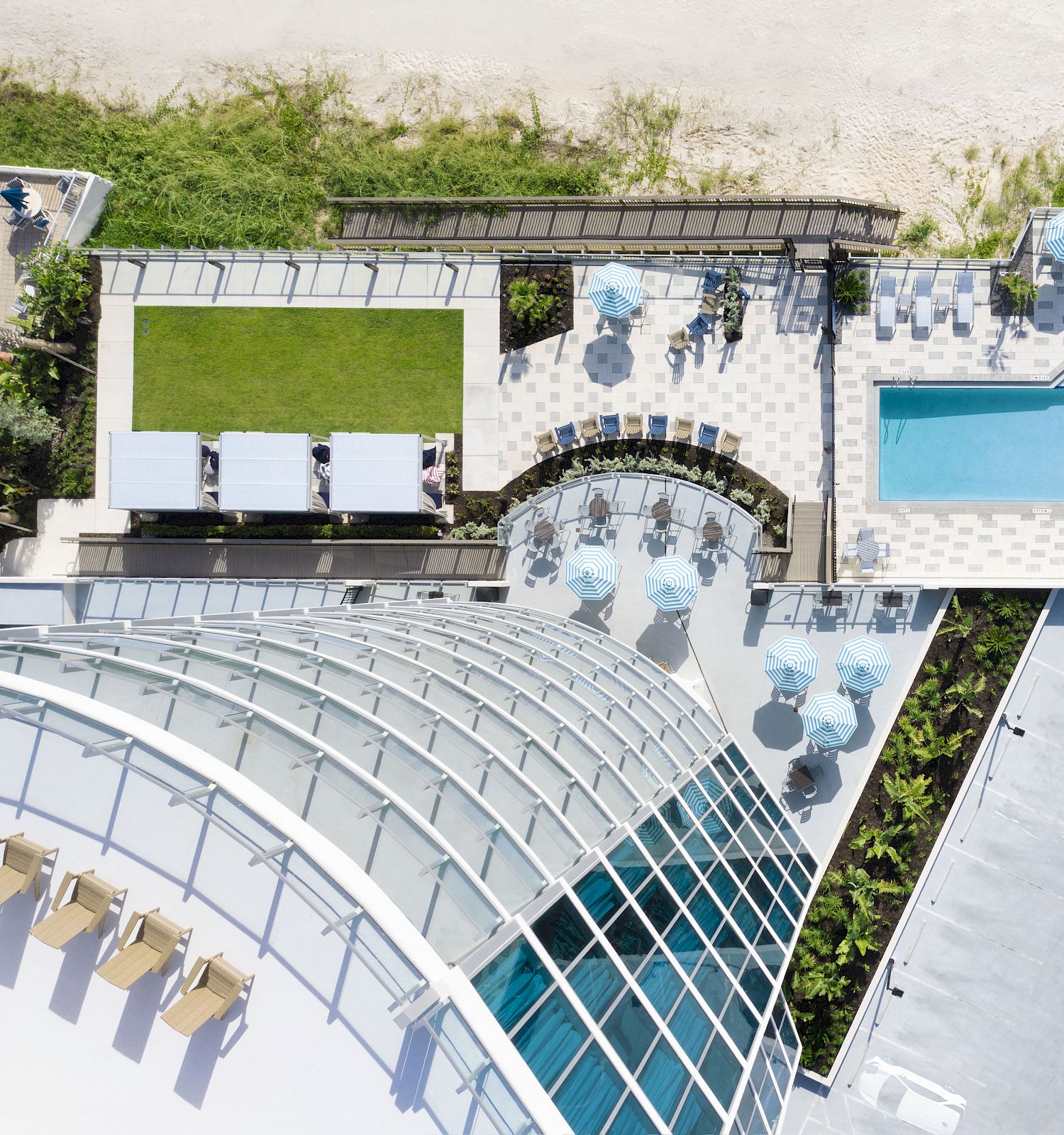 An aerial view of a building with a domed glass structure, adjacent pool area, lounge chairs, umbrellas, and a grassy section near a sandy beach.