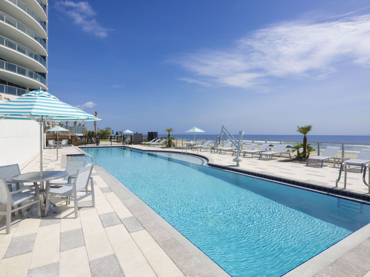 A luxurious outdoor pool area with lounge chairs, umbrellas, tables, and a stunning ocean view on a sunny day.