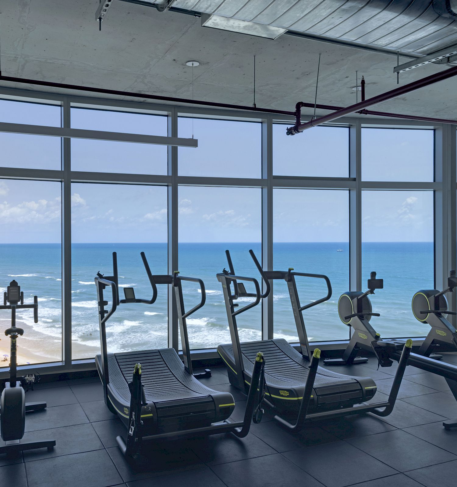 A gym with treadmills and exercise bikes overlooks a scenic beach and ocean through large windows, providing a stunning workout view.