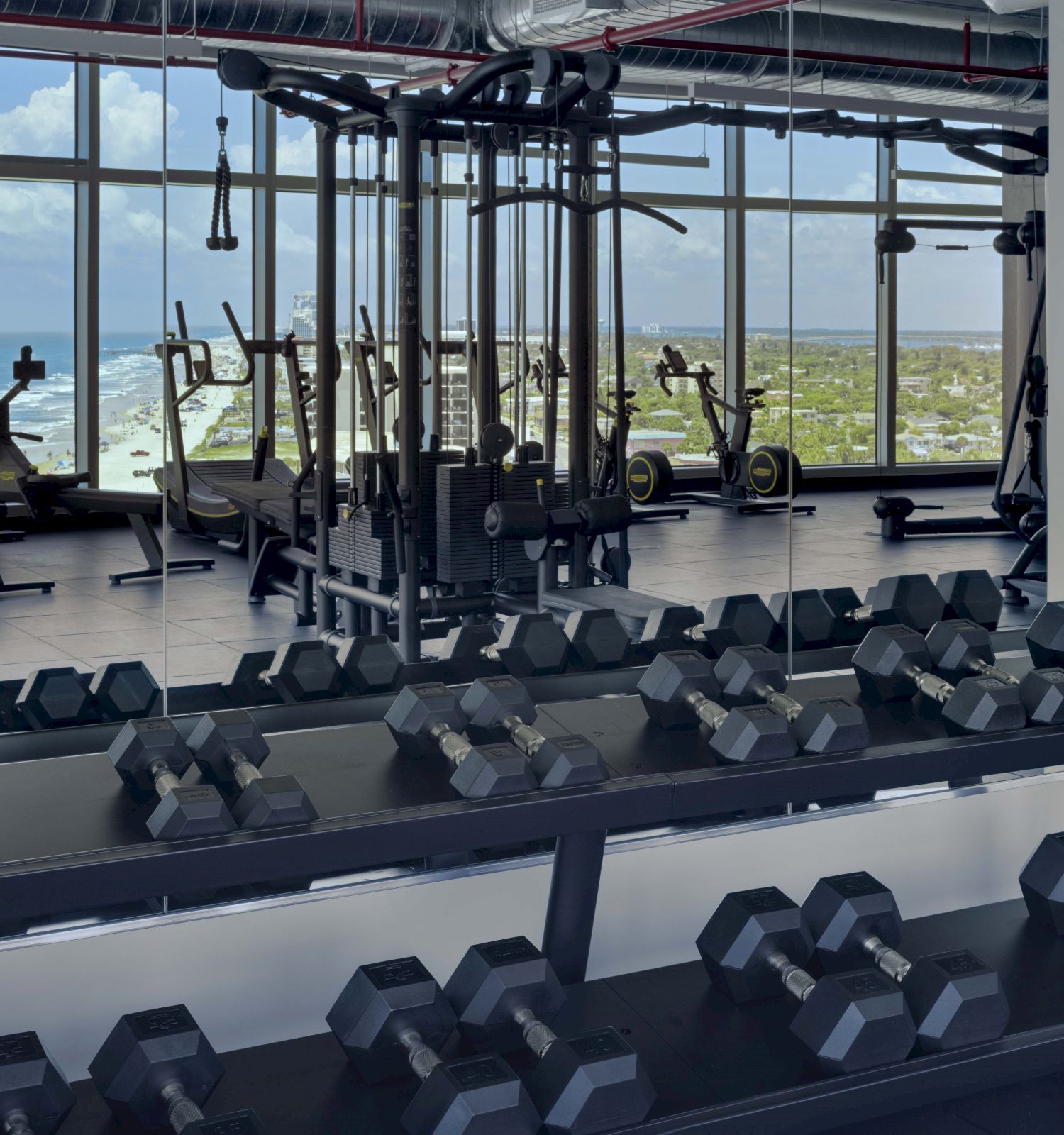 A gym with dumbbells, various weight machines, and rowing machines by large windows overlooking a scenic view of the ocean and coastline.