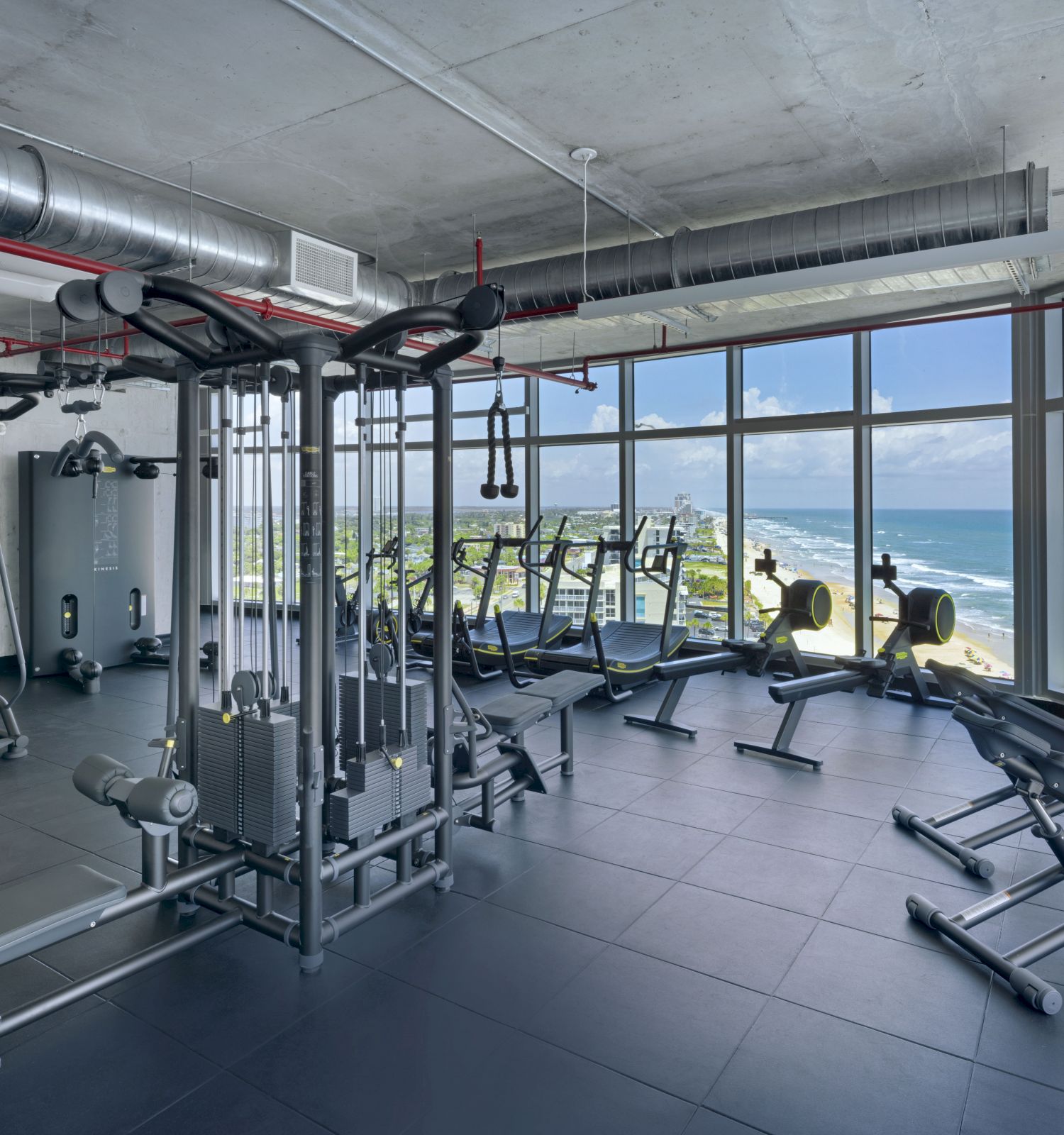 A modern gym with various exercise equipment is shown, featuring large windows with a scenic ocean view, creating a bright, motivating atmosphere.