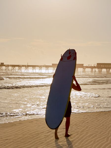 A person is standing on the beach holding a surfboard, facing the ocean with a pier visible in the background against the setting sun.