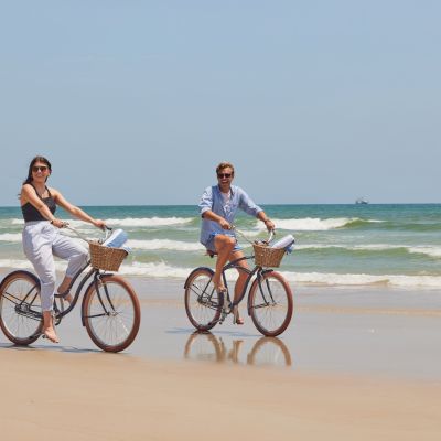 Two people are riding bicycles with baskets along a sandy beach with waves in the background, under a clear sky, enjoying a sunny day.