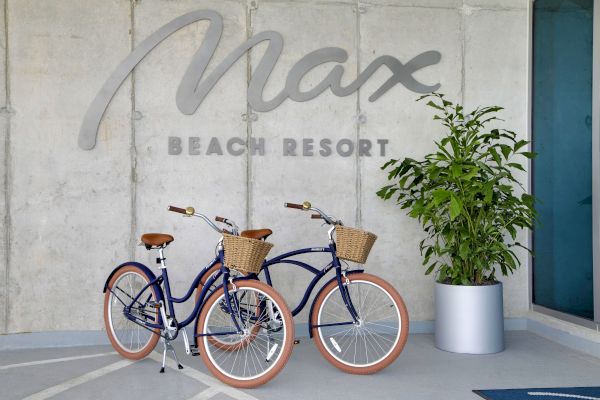 Two blue bicycles with brown seats and wicker baskets are parked in front of a wall with "Max Beach Resort" signage, alongside a potted plant.