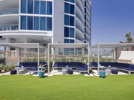 The image shows an outdoor lounge area with canopied seating, blue cushions, and small tables on a green lawn beside a modern building.