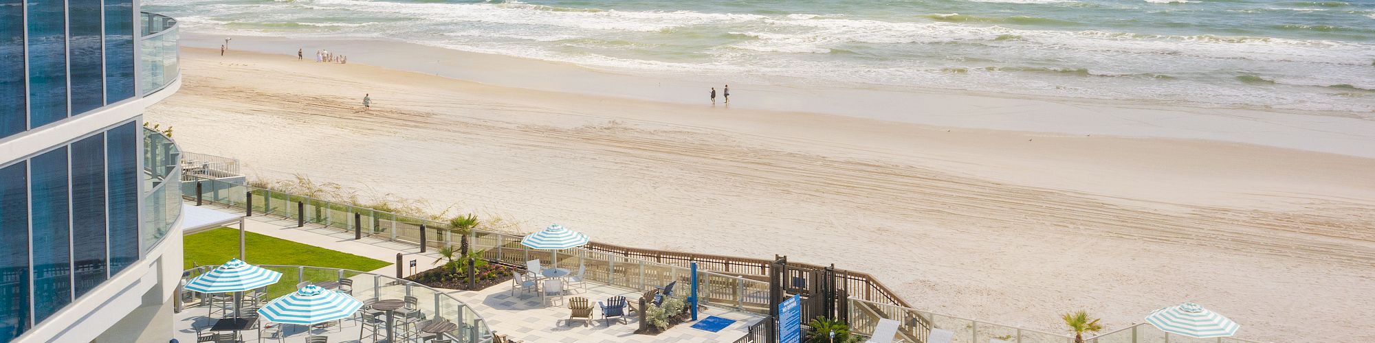 This image shows a beachfront scene with a swimming pool, sun loungers, and striped umbrellas, next to a beach with waves and a few people walking.