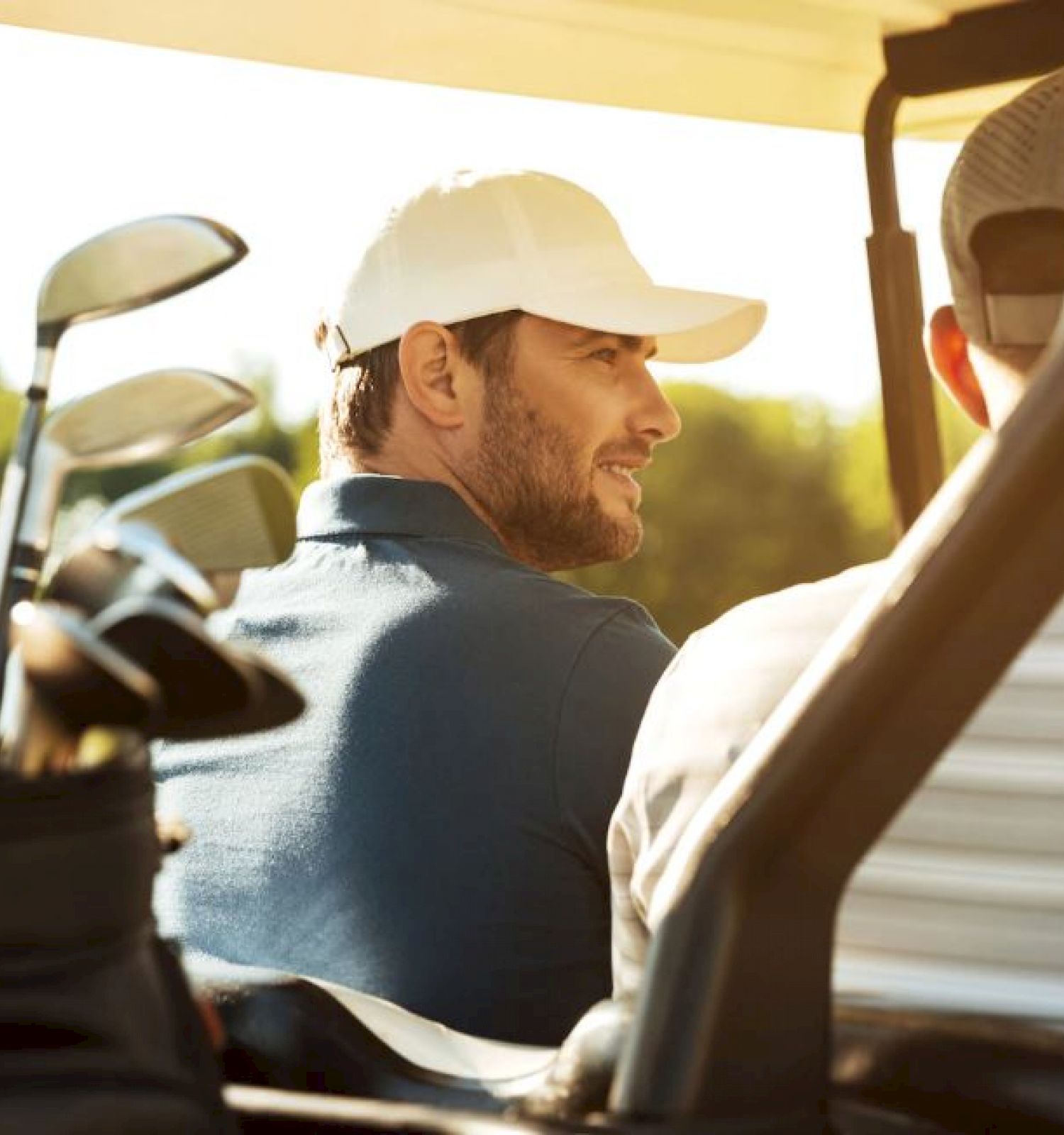 Two men ride in a golf cart, one wearing a white cap, the other a striped shirt. Golf clubs are visible behind them, with greenery in the background.