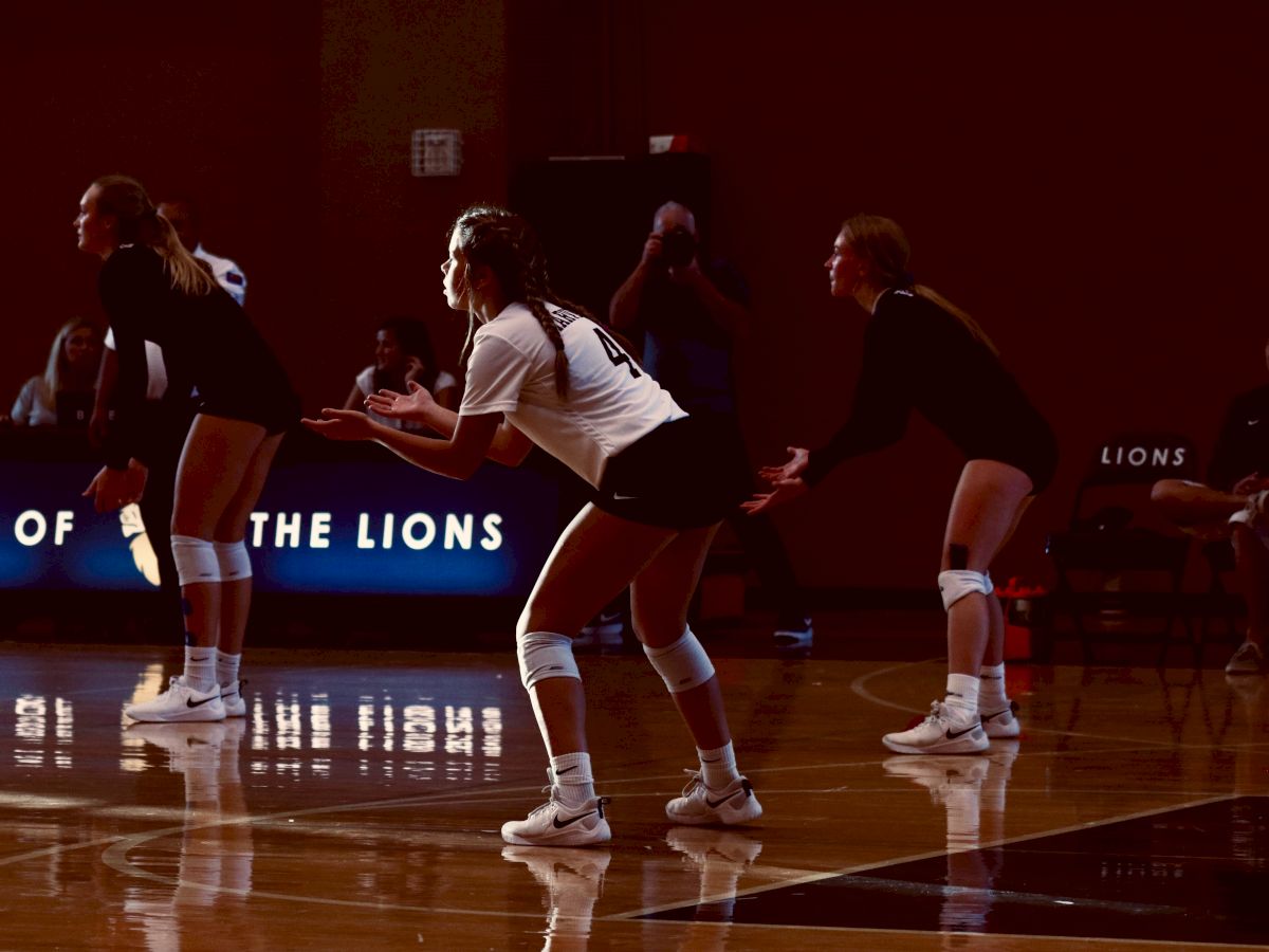 Three volleyball players in ready stance on the court, with a sign in the background reading "Home of the Lions."