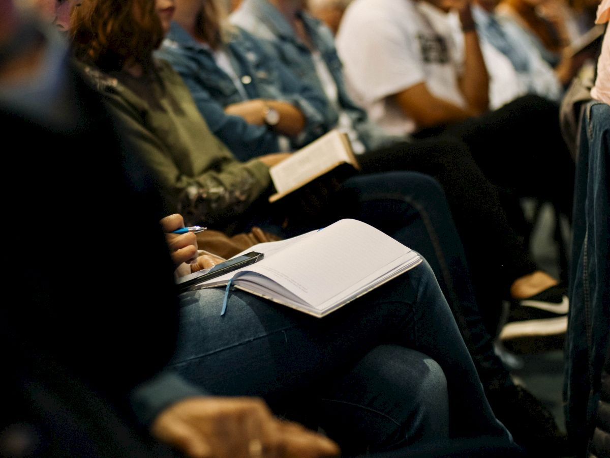 The image shows people seated closely together, some holding books and notebooks, appearing to be engaged in an event or lecture.