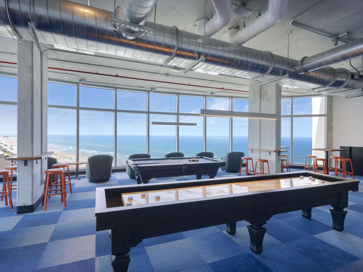 A modern room with large windows showing a coastal view, featuring a pool table, shuffleboard table, and seating area with high stools and armchairs.