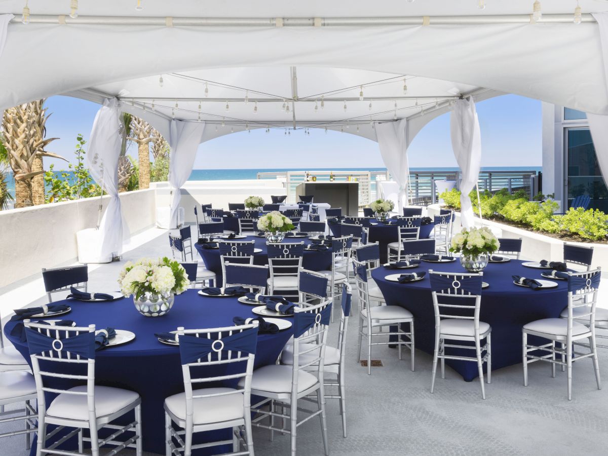 The image shows a beautifully arranged outdoor event setup, with round tables covered in navy blue tablecloths and white chairs, under a white canopy near the ocean.