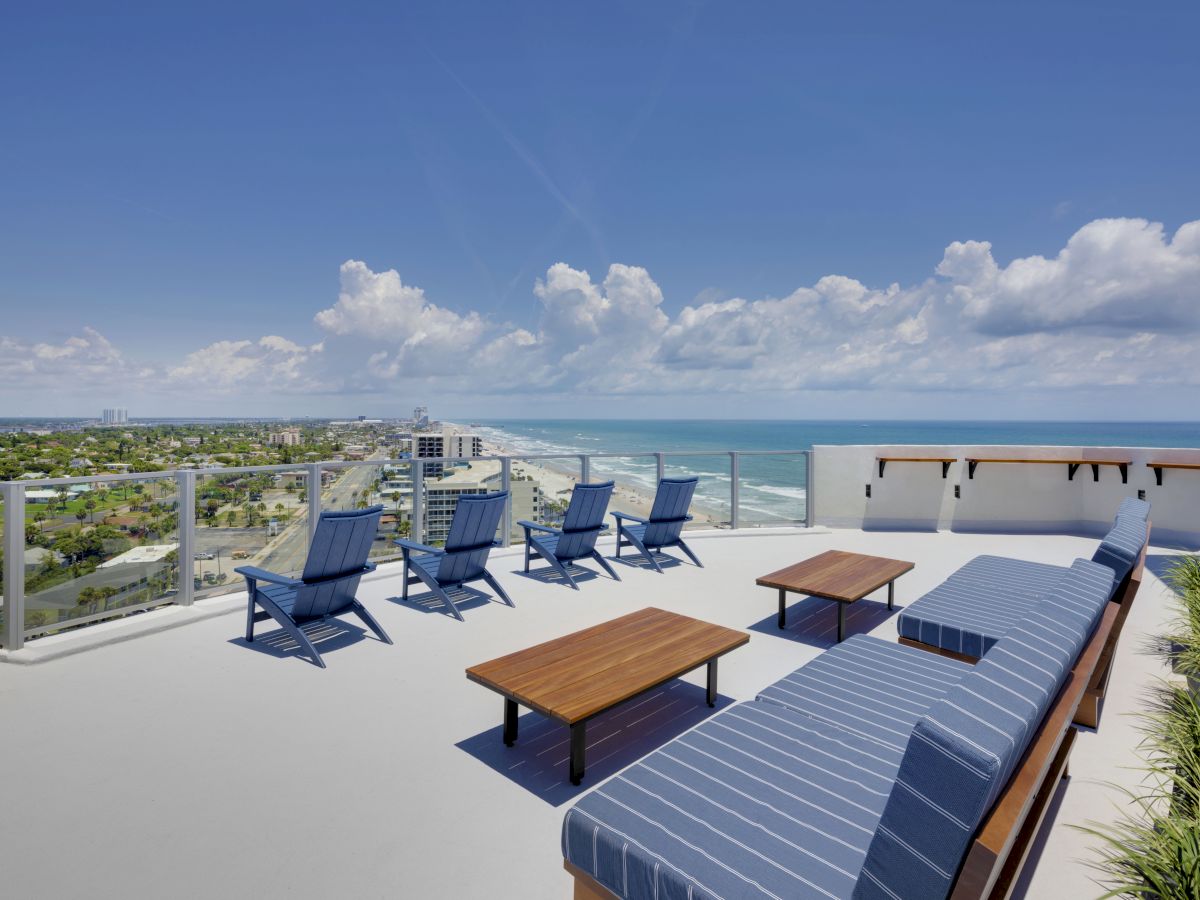 The image shows a rooftop terrace with lounge chairs and tables overlooking a coastal cityscape and the ocean under a blue sky with clouds.