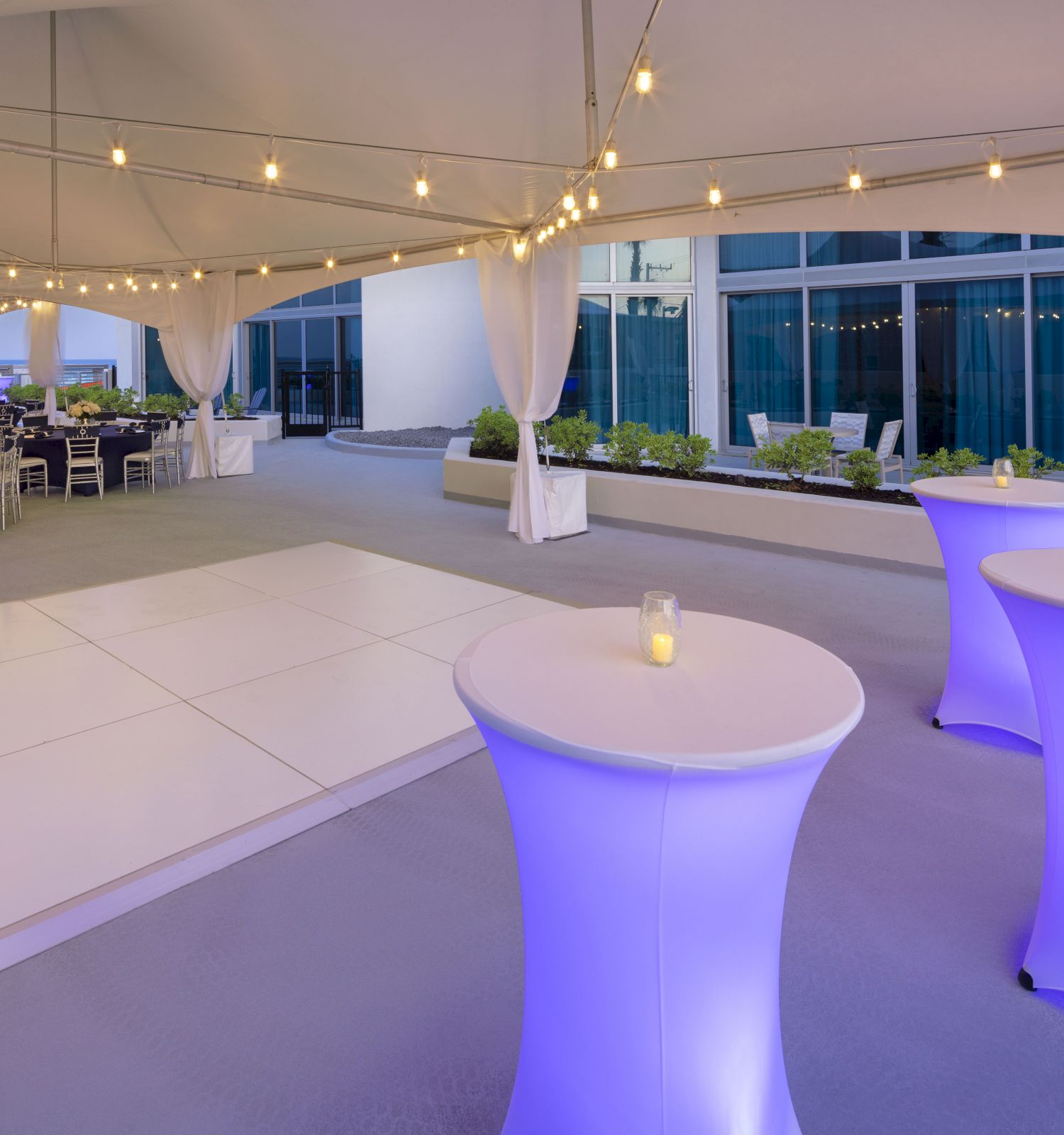 An elegant outdoor event setup with high tables lit in blue, a dance floor, string lights overhead, and tables with chairs in the background.