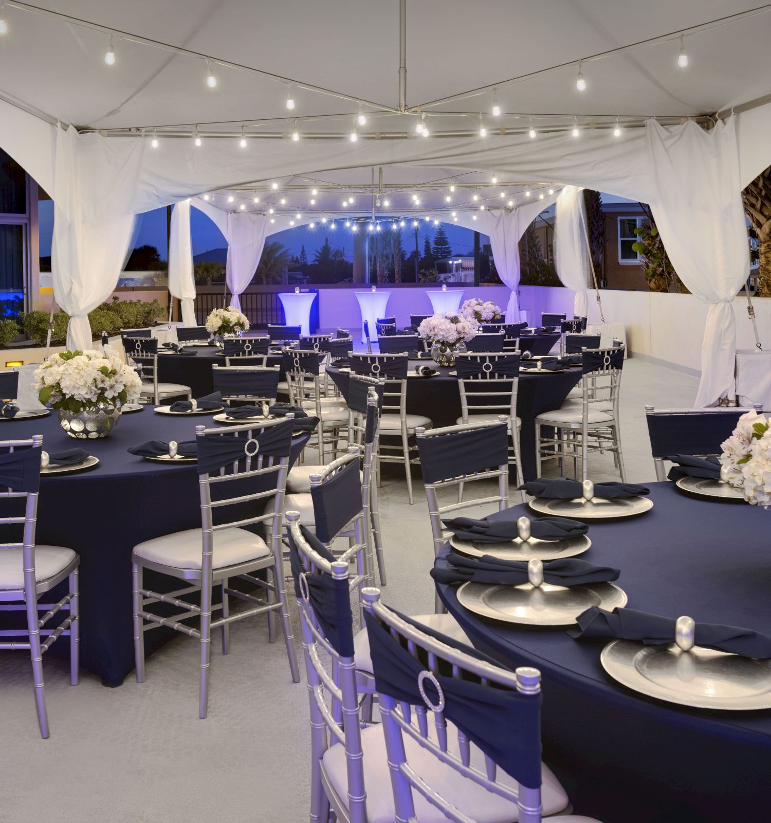 An elegant outdoor event setup with round tables and black tablecloths, silver chairs, floral centerpieces, and string lights under a tented canopy.