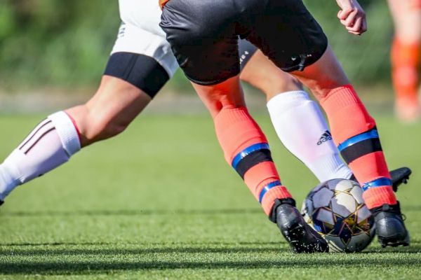 Two soccer players in action, one in orange socks and the other in white, compete for the ball on a green field.