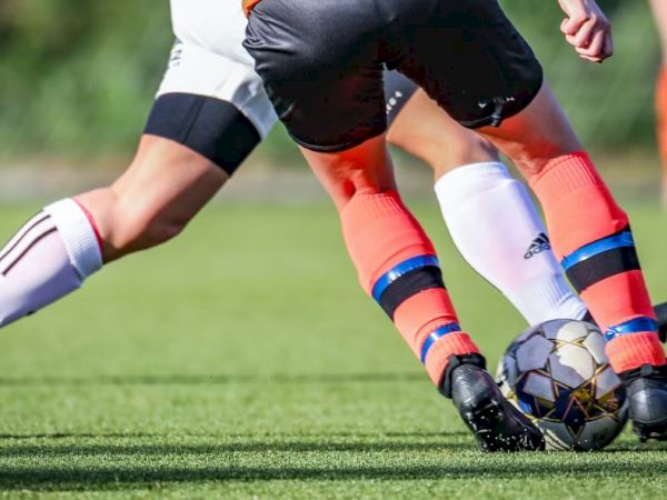 Two soccer players in action, one in orange socks and the other in white, compete for the ball on a green field.