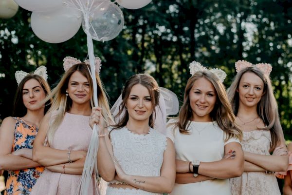 A group of five women, wearing dresses and cat ear headbands, pose outdoors with crossed arms and balloons, smiling confidently.